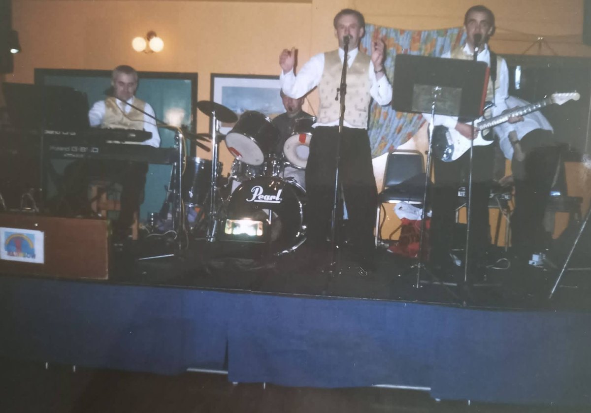 The family wedding band on stage in Ireland in matching gold waistcoats: keys, drums, a singer, and Colin on electric guitar at the far right