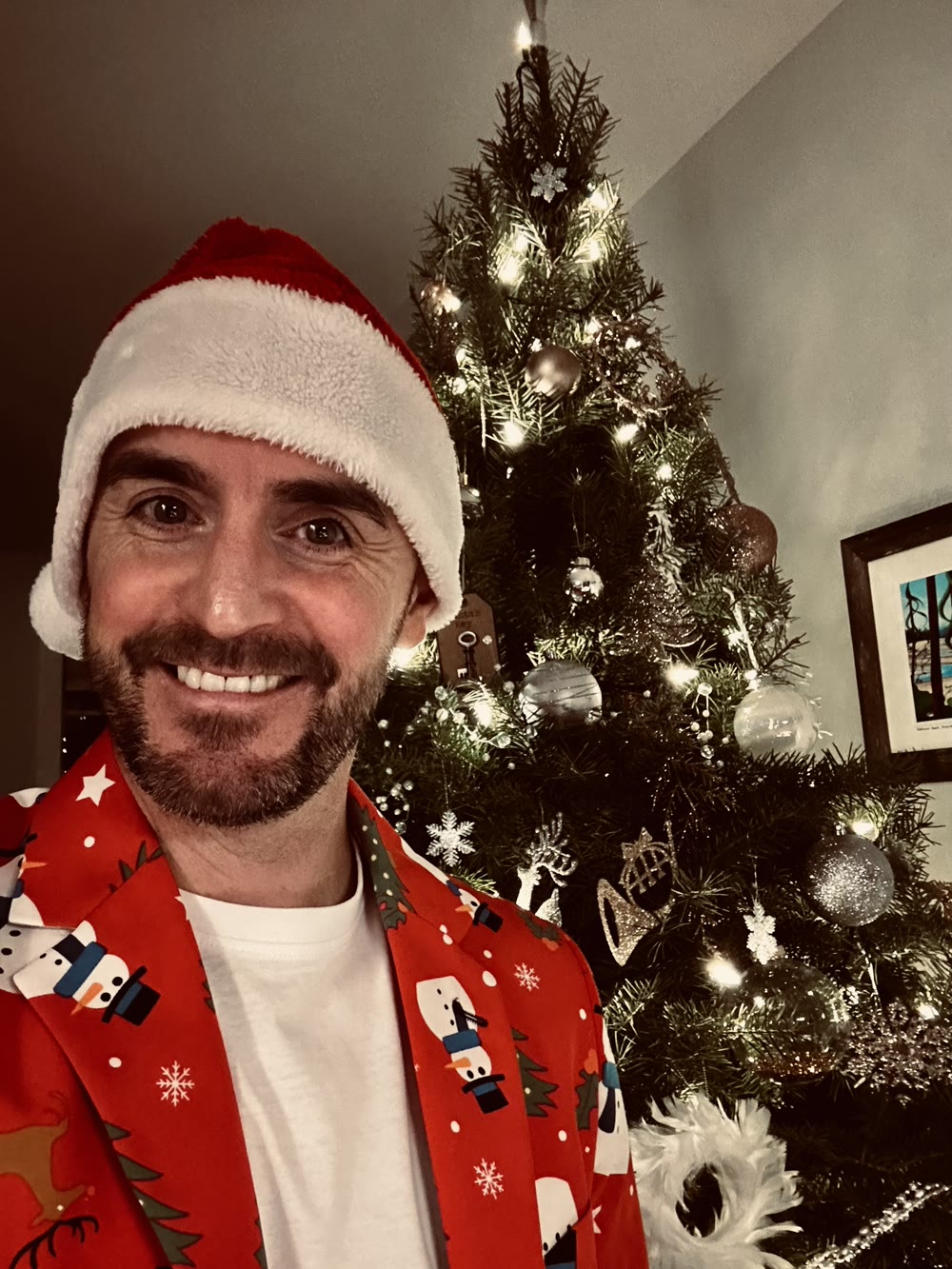 Colin in a Santa hat and a red snowman blazer, smiling in front of a lit Christmas tree